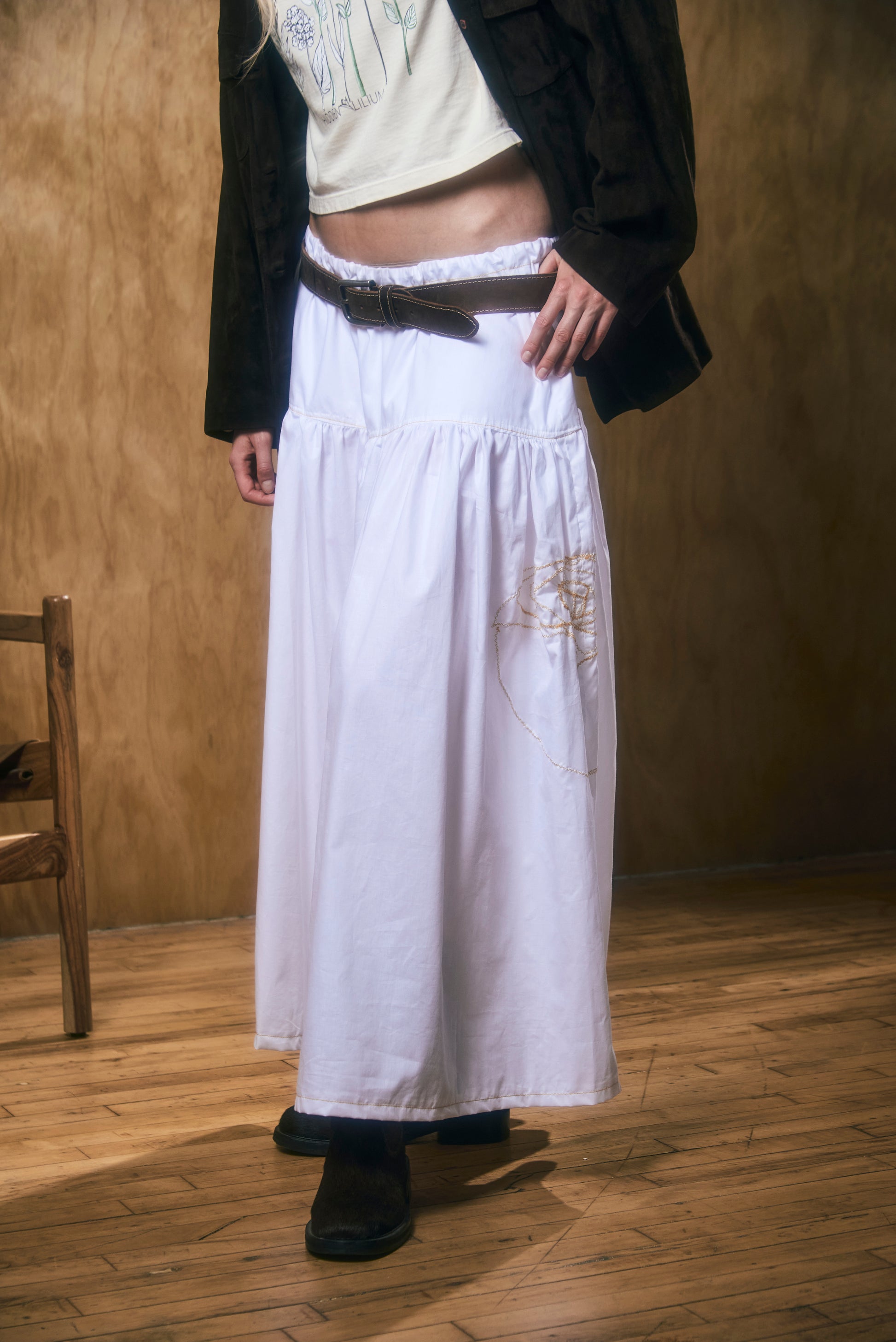 Person wearing a long white skirt with floral embroidery against a wooden floor and wall.
