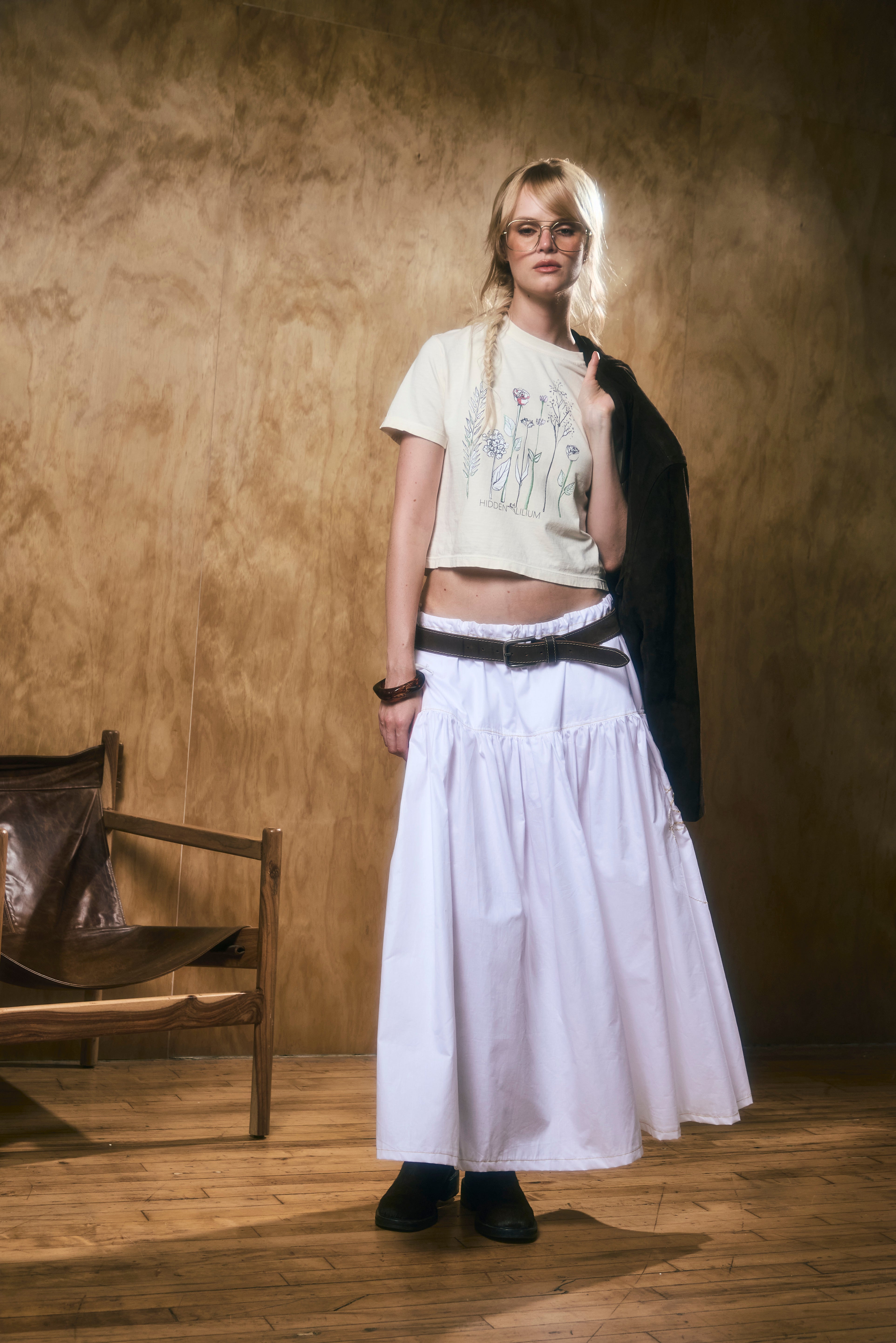 woman wearing a light yellow t-shirt and long white skirt standing against a wooden wall.