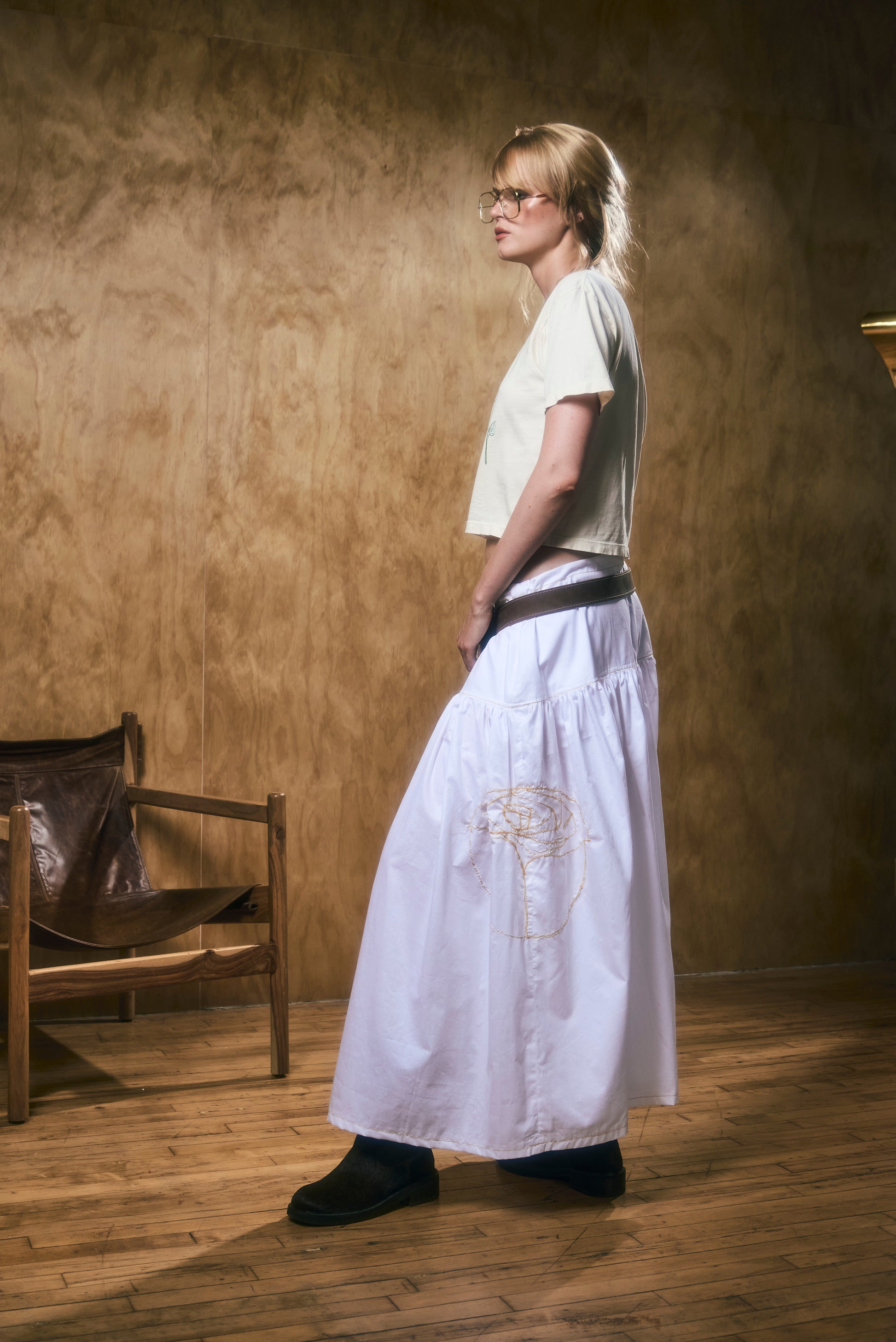 A woman wearing a long white skirt with flower embroidery in a room with wooden floor and walls.