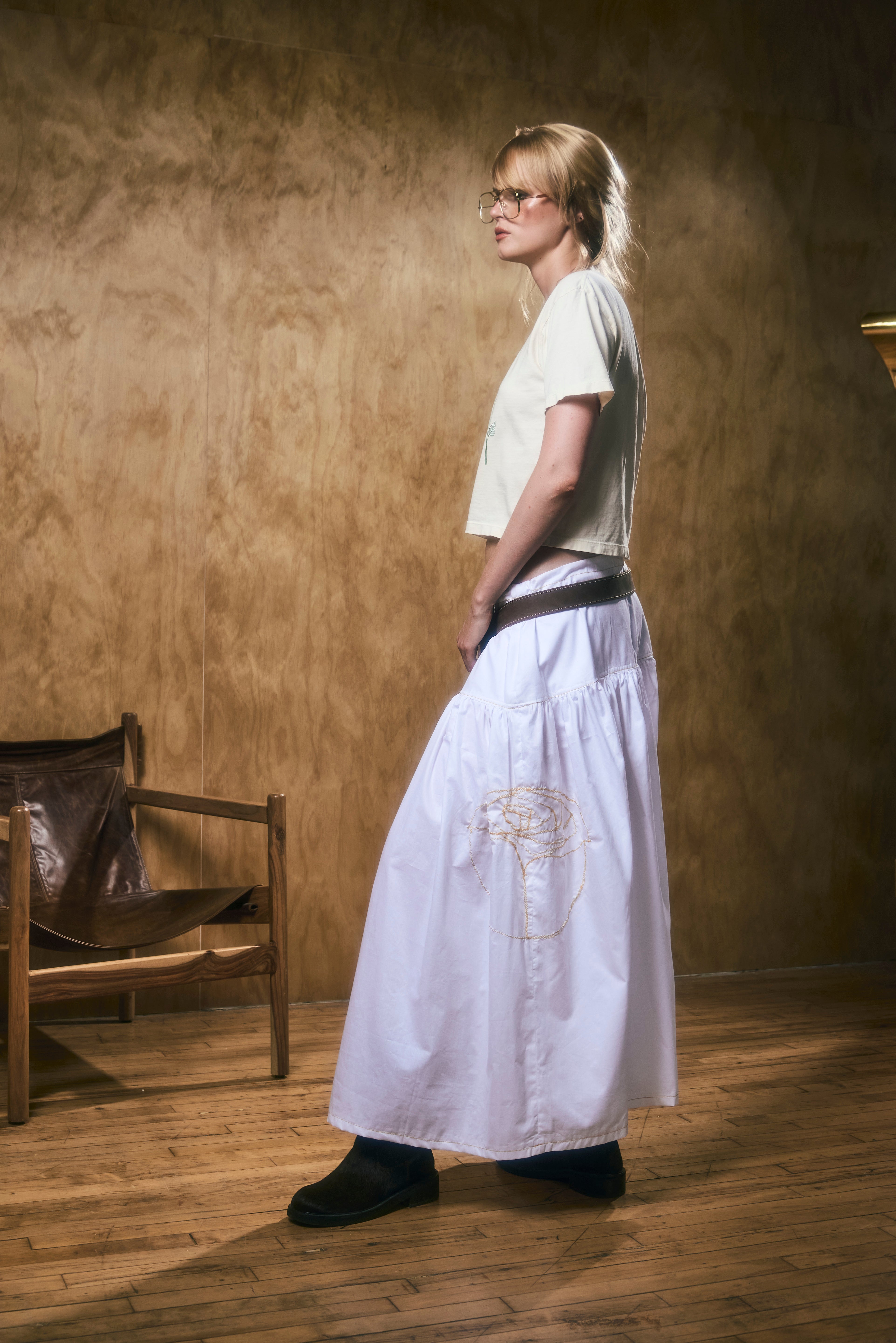 A woman wearing a long white skirt with flower embroidery in a room with wooden floor and walls.