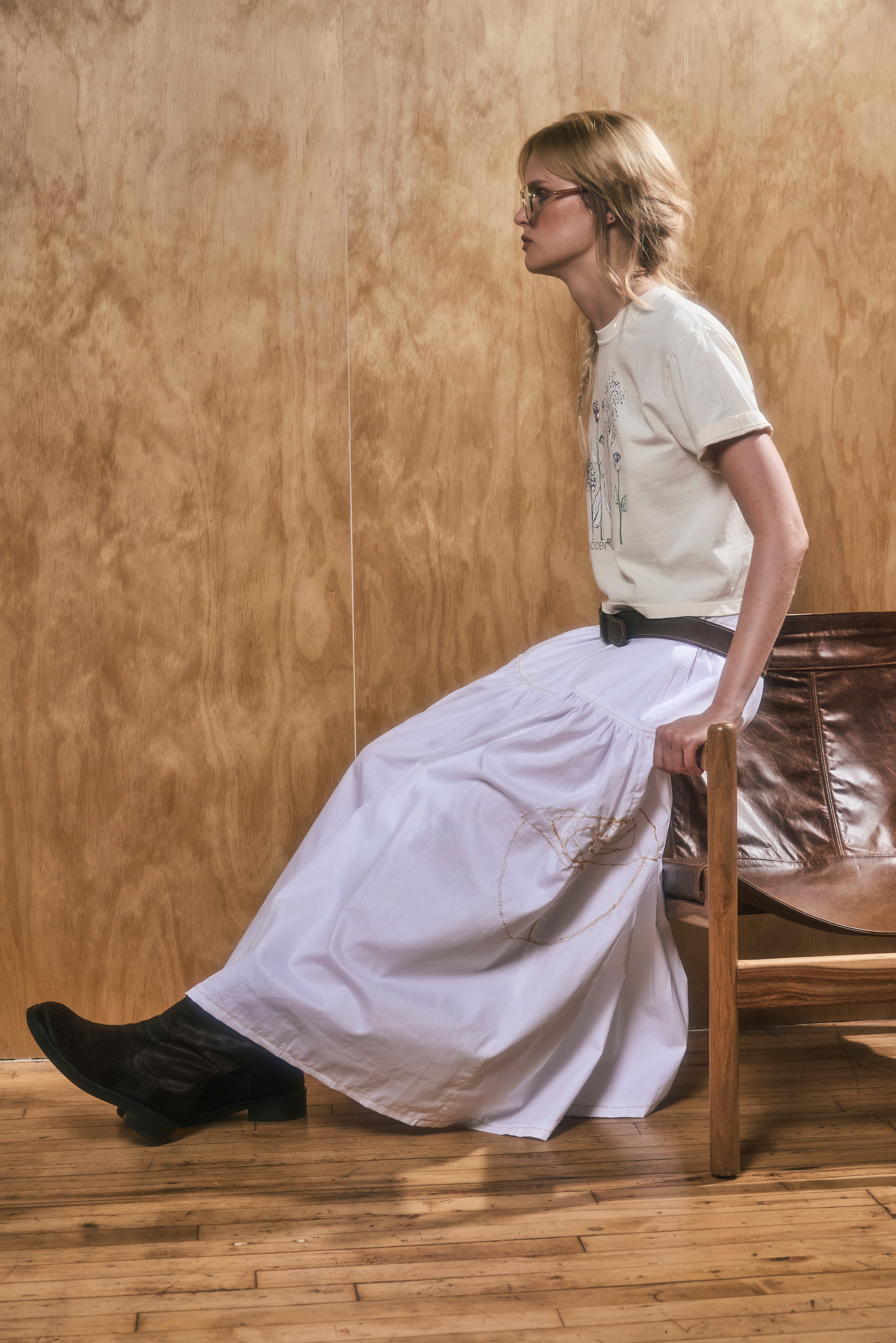 women sitting on a chair wearing a white skirt, a light yellow t-shirt, and black shoes in a wooden room.