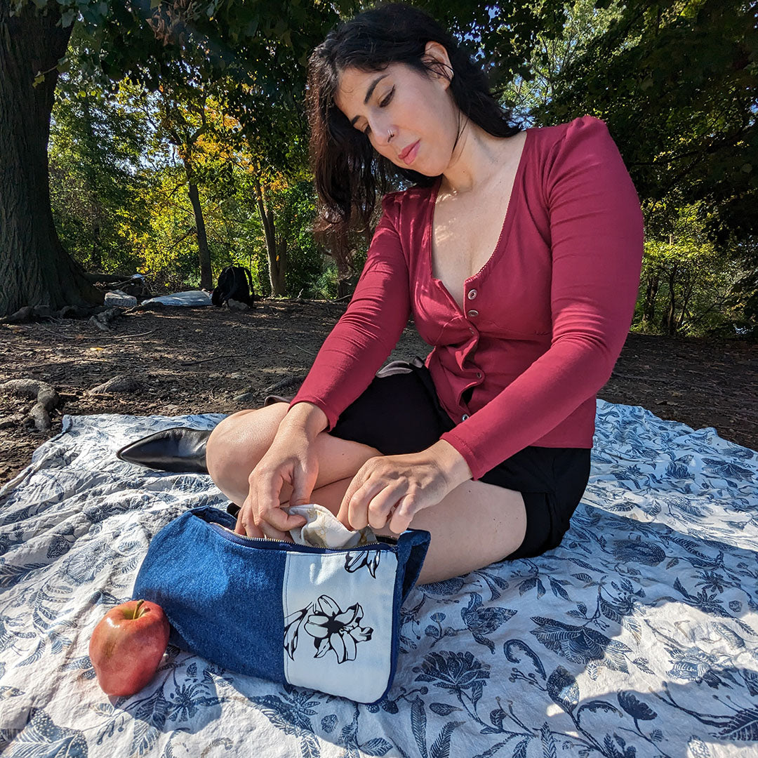 Woman sitting on a blanket in a park, wearing a red-burgundy shirt, opening a blue bag.