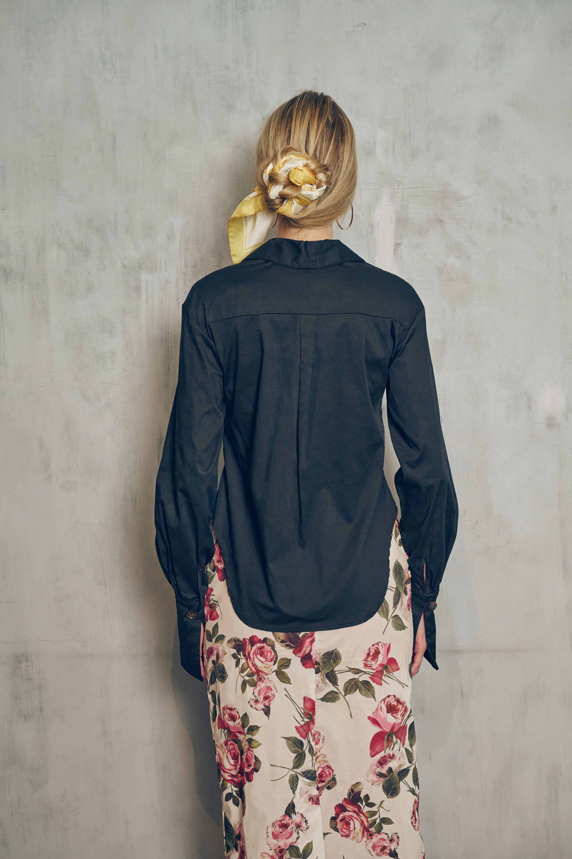 Back view of a person wearing a black shirt and a floral skirt against a gray wall