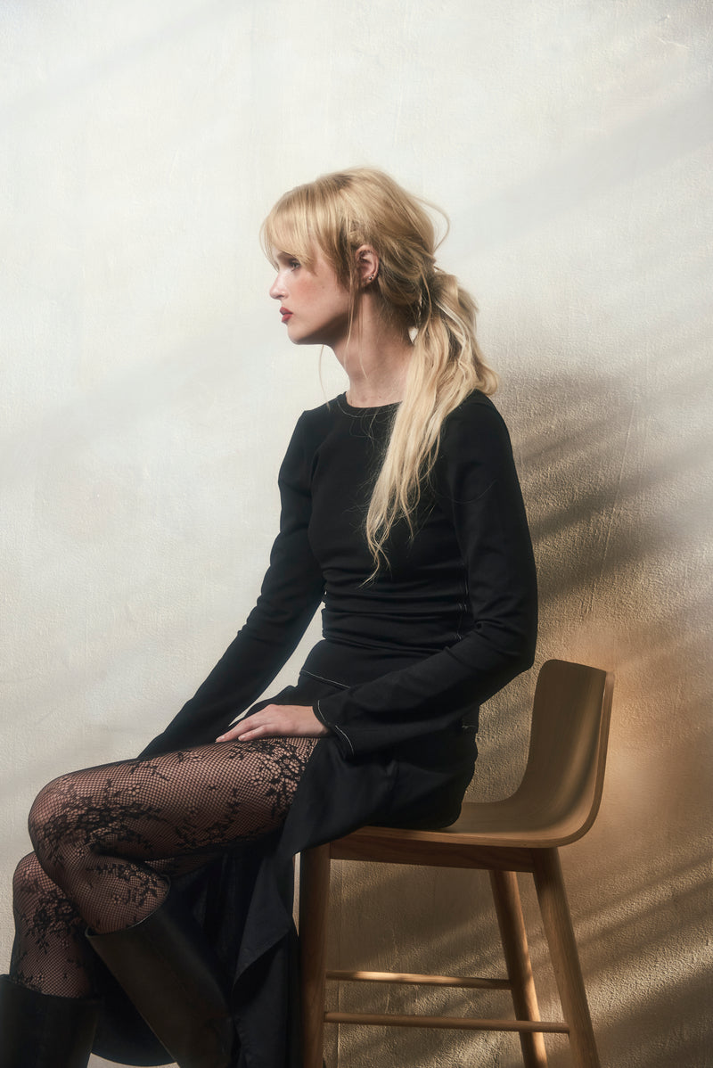 Woman wearing a black long-sleeved shirt and black skirt, sitting on a wooden stool against a textured wall.