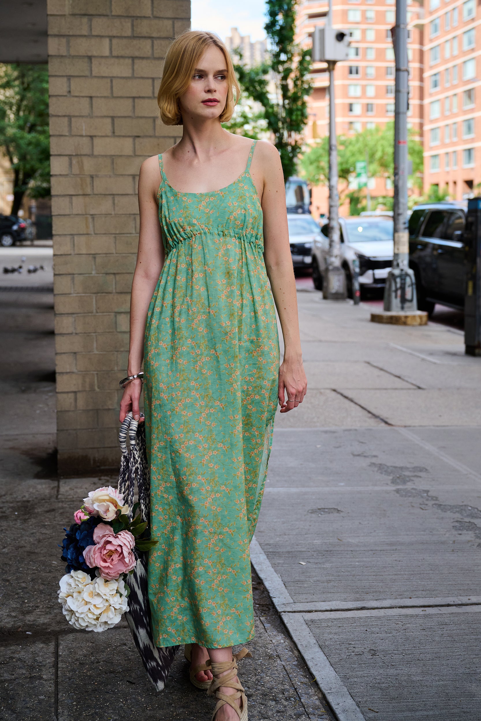 Woman in a green floral dress holding flowers on a city street.