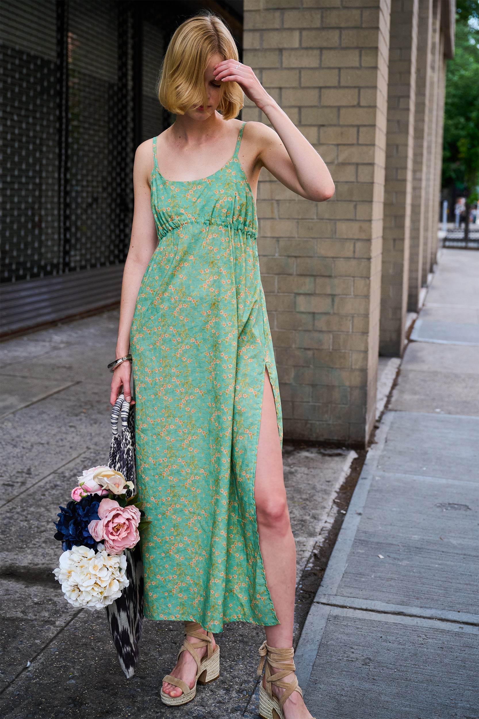 Woman in a green floral dress standing on a sidewalk holding flowers.