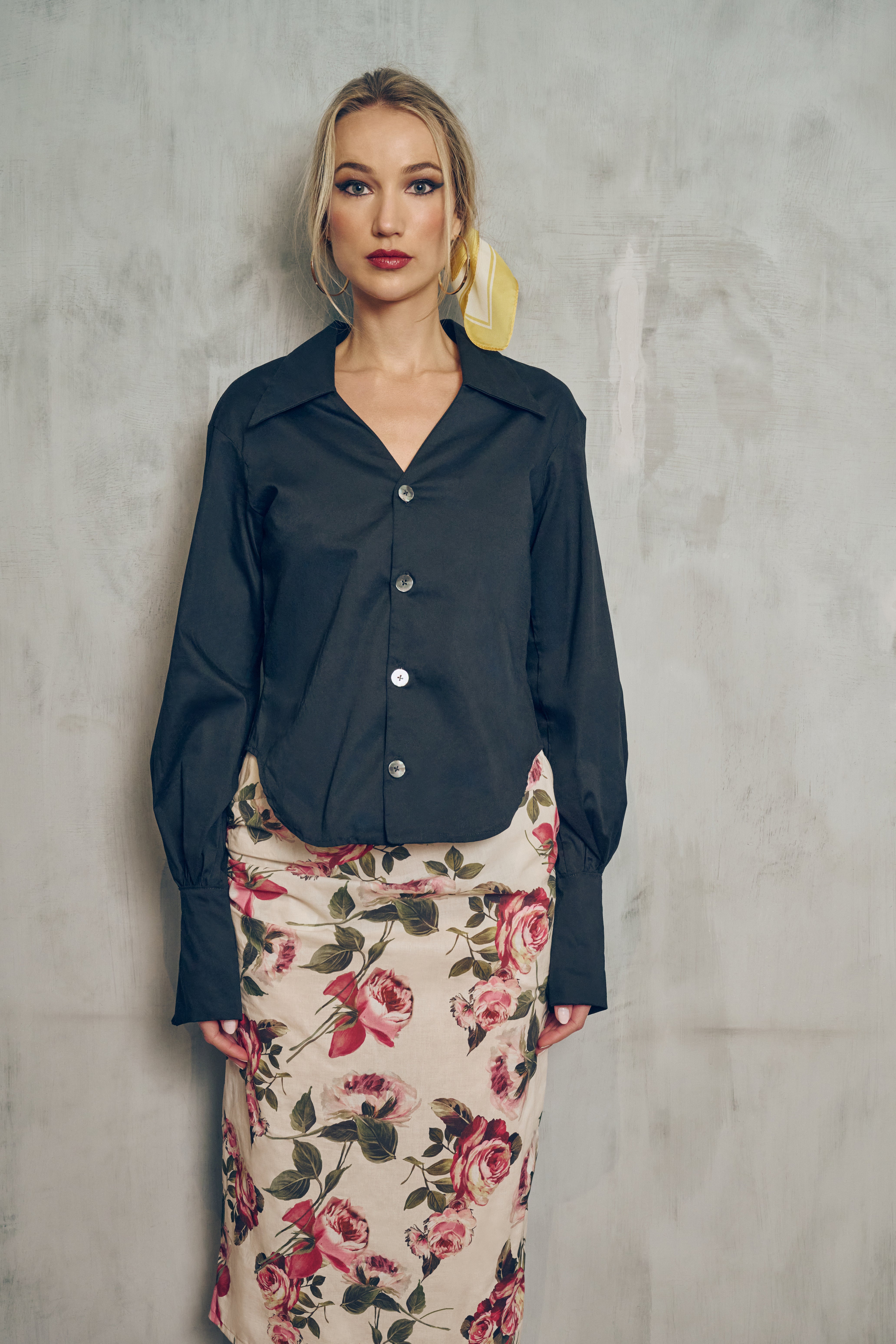  Front view of a person wearing a black button-down shirt with a V-neck line and big cuffs  and a floral skirt against a gray wall
