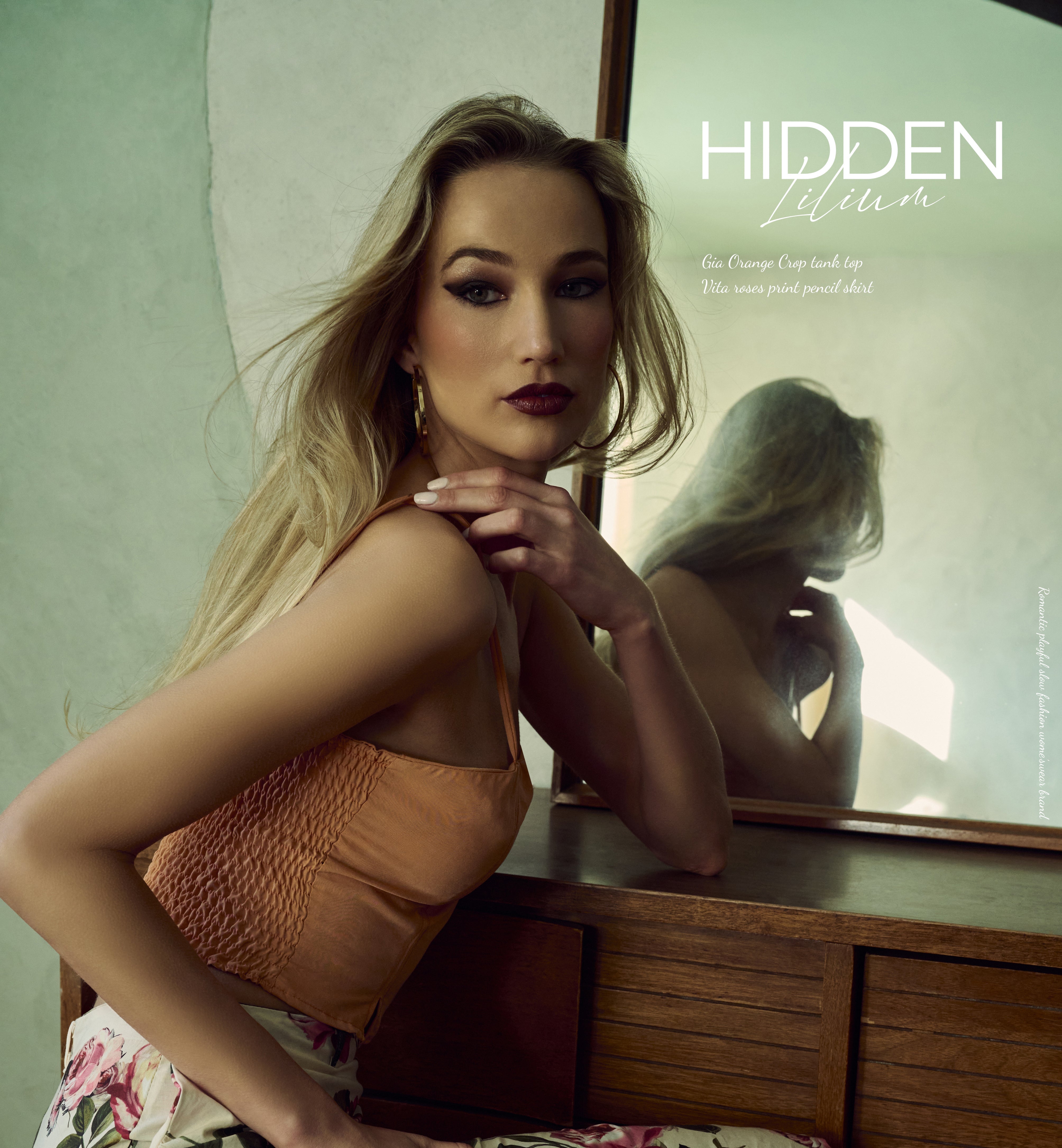 Woman in an orange tank and floral skirt sitting on a wooden chair with a mirror in the background, featuring the brand 'Hidden Lilium'