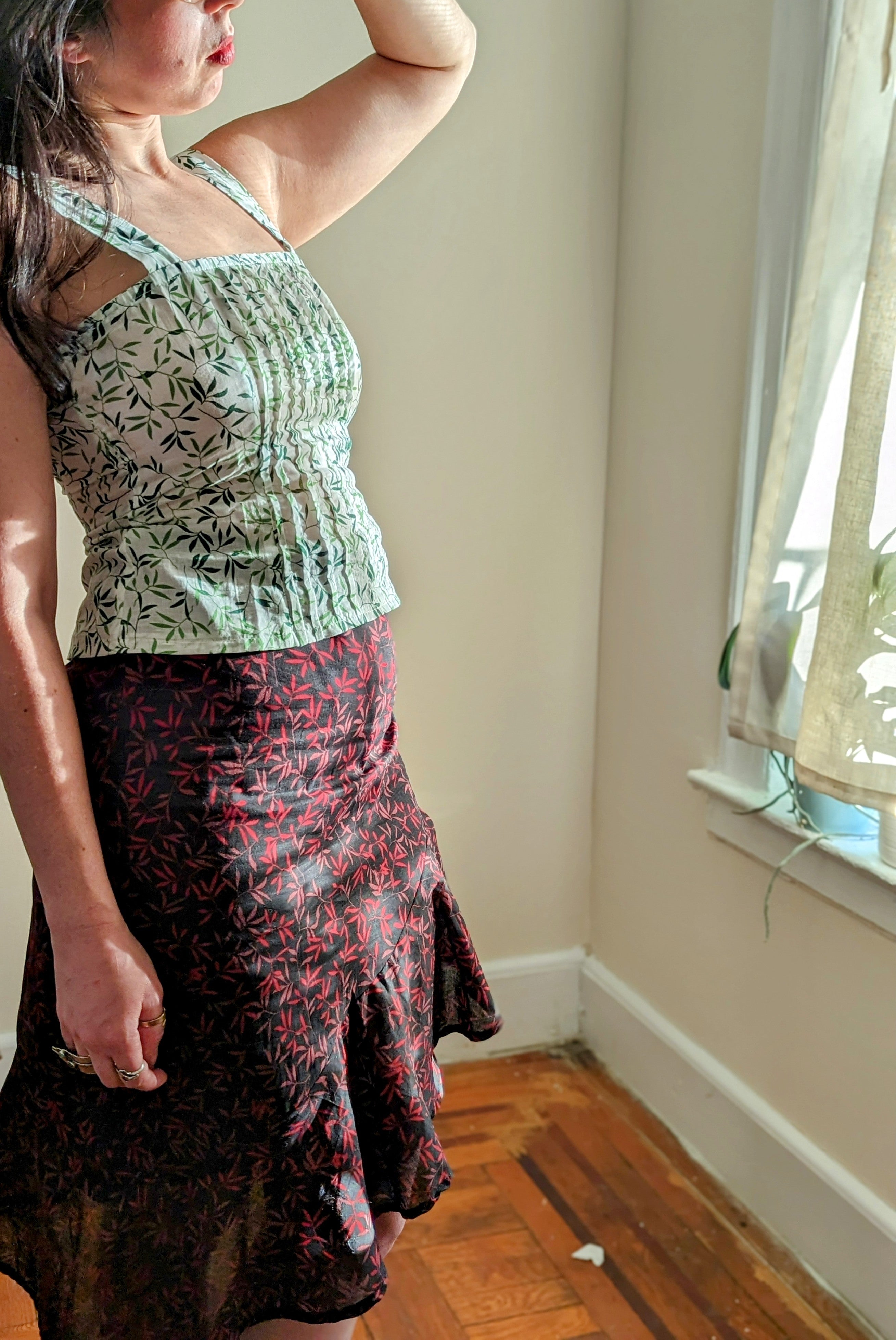 Woman wearing a green leaf patterned top and black floral skirt in a room with a window.