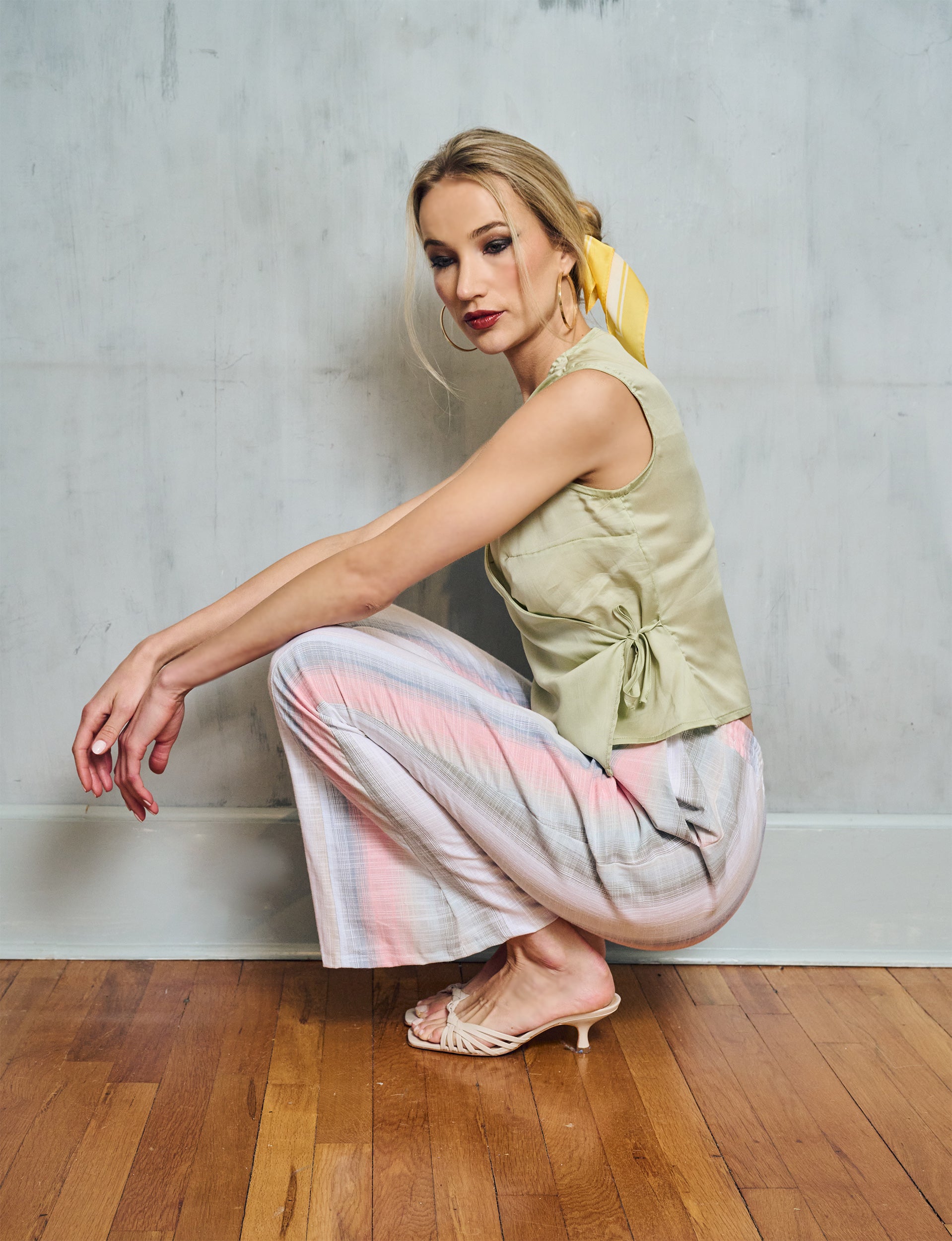 Woman sitting on the floor wearing a light green sleeveless top and colorful pants against a plain wall.