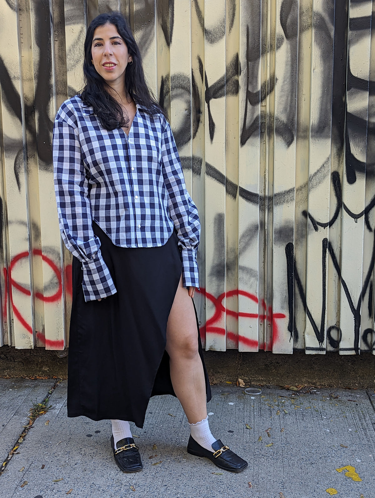 A woman stands outdoors in front of a graffiti-covered corrugated metal wall, wearing a blue and white gingham button-up shirt, a long black skirt, white socks, and black loafers with gold hardware.