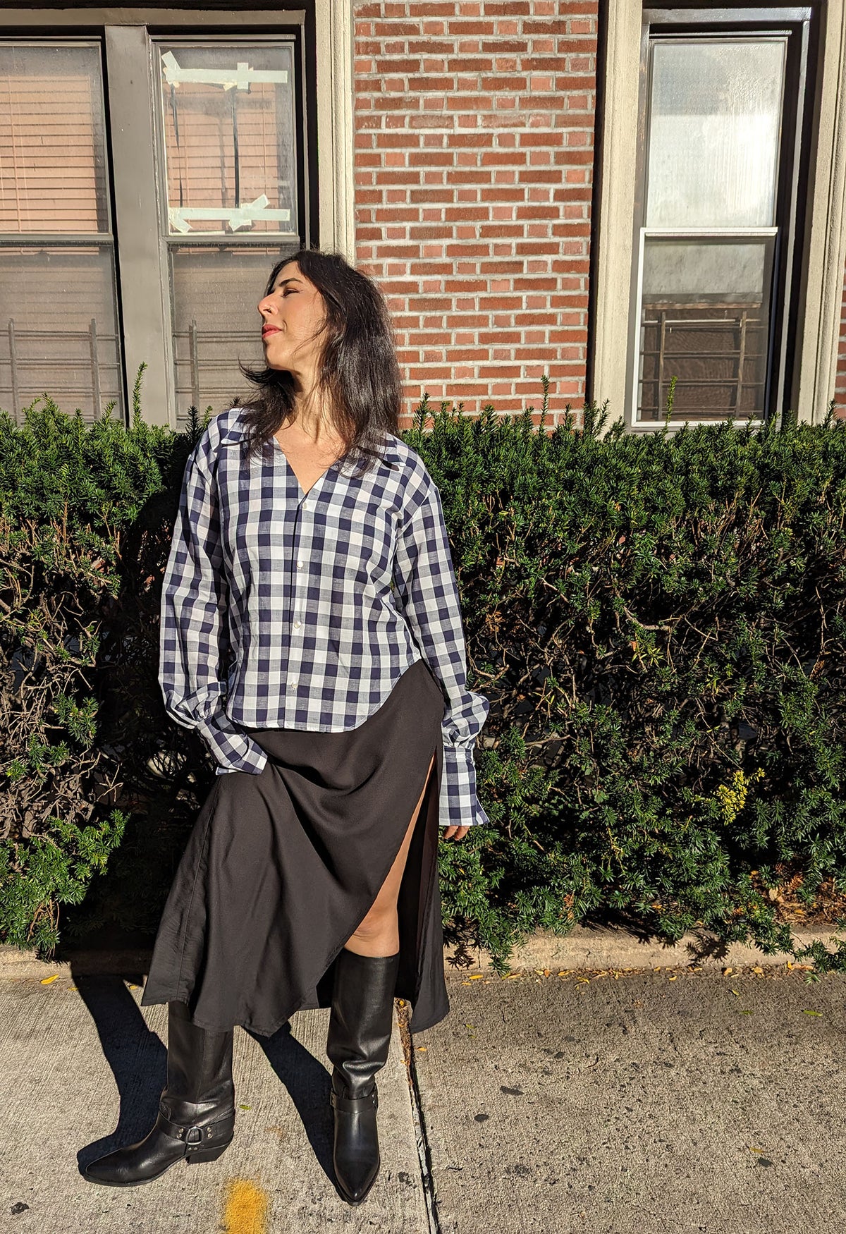 A woman stands outdoors in front of a brick building and green hedge, wearing a Navy and white gingham button-up shirt, a black slit skirt, and black knee-high boots.