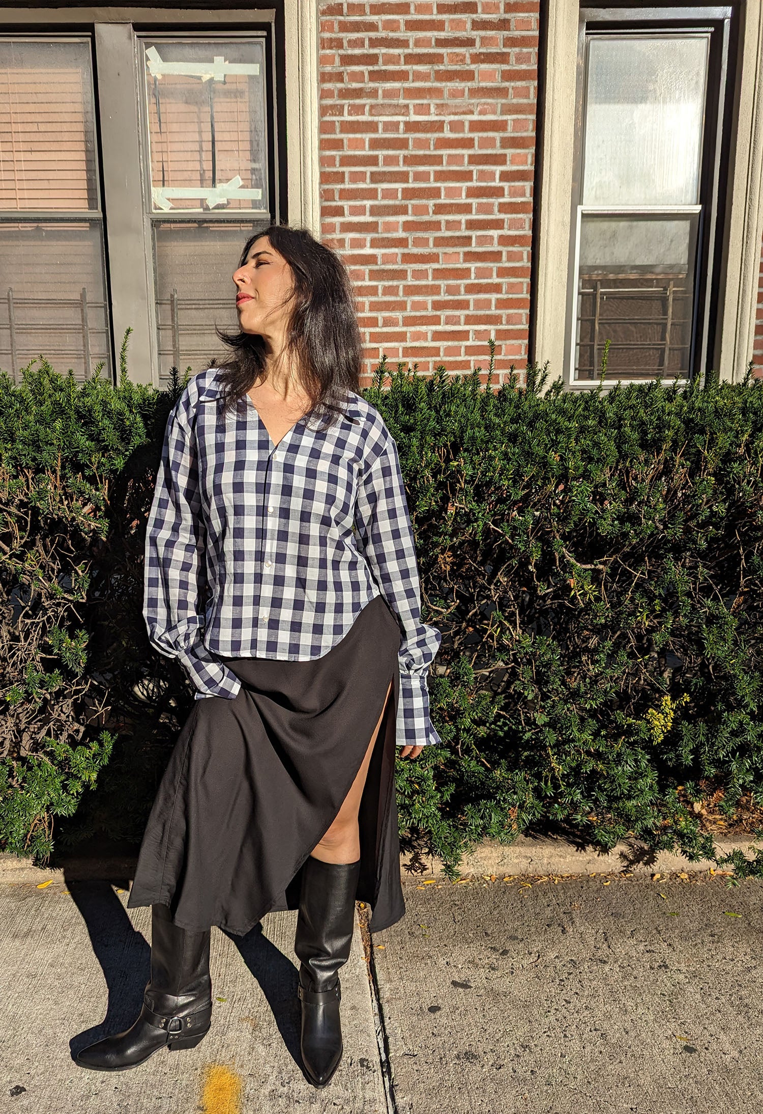 A woman stands outdoors in front of a brick building and green hedge, wearing a Navy and white gingham button-up shirt, a black slit skirt, and black knee-high boots.