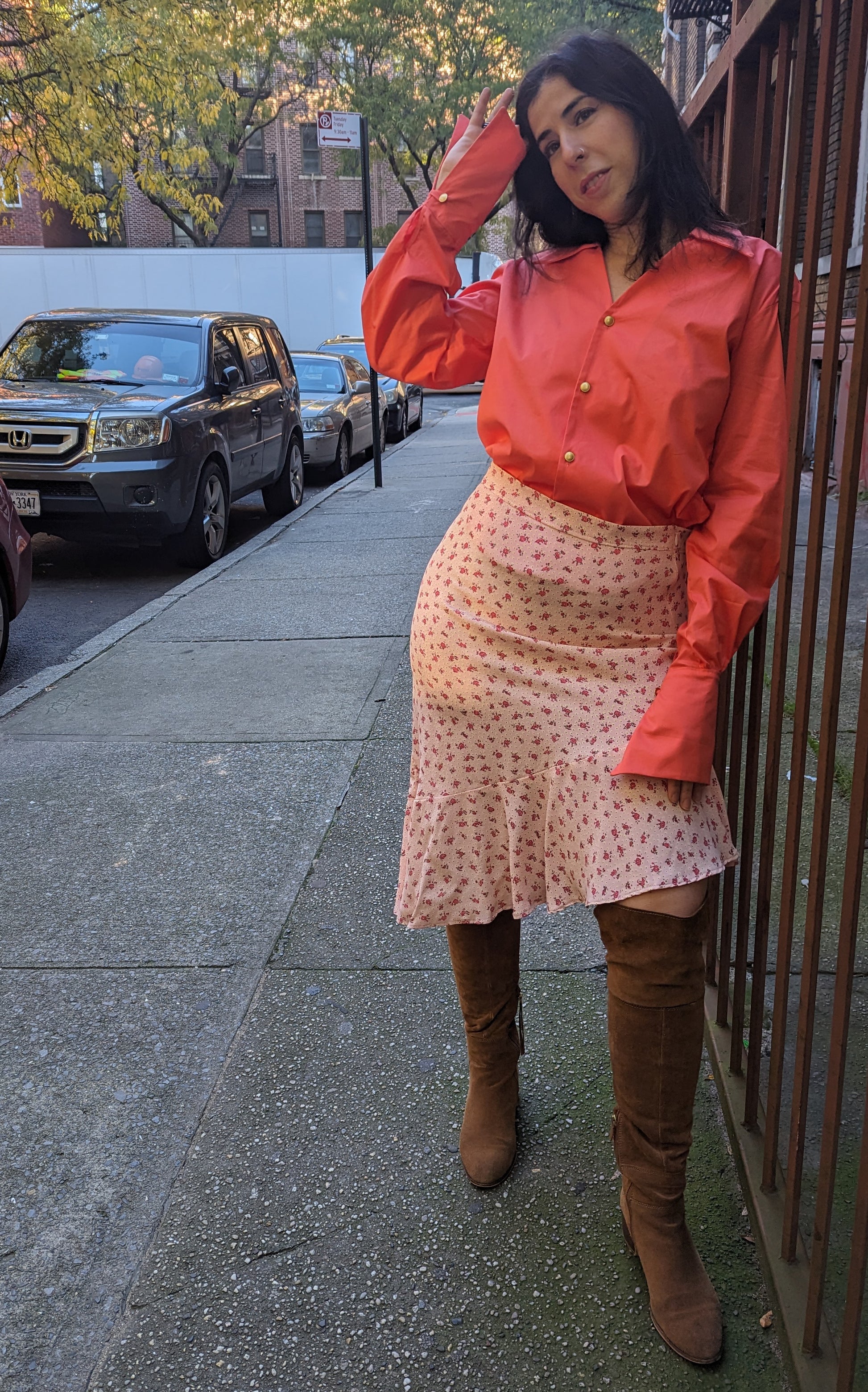 Woman in a  ornge button down shirt and a floral pink skirt standing on a sidewalk with cars and trees in the background.