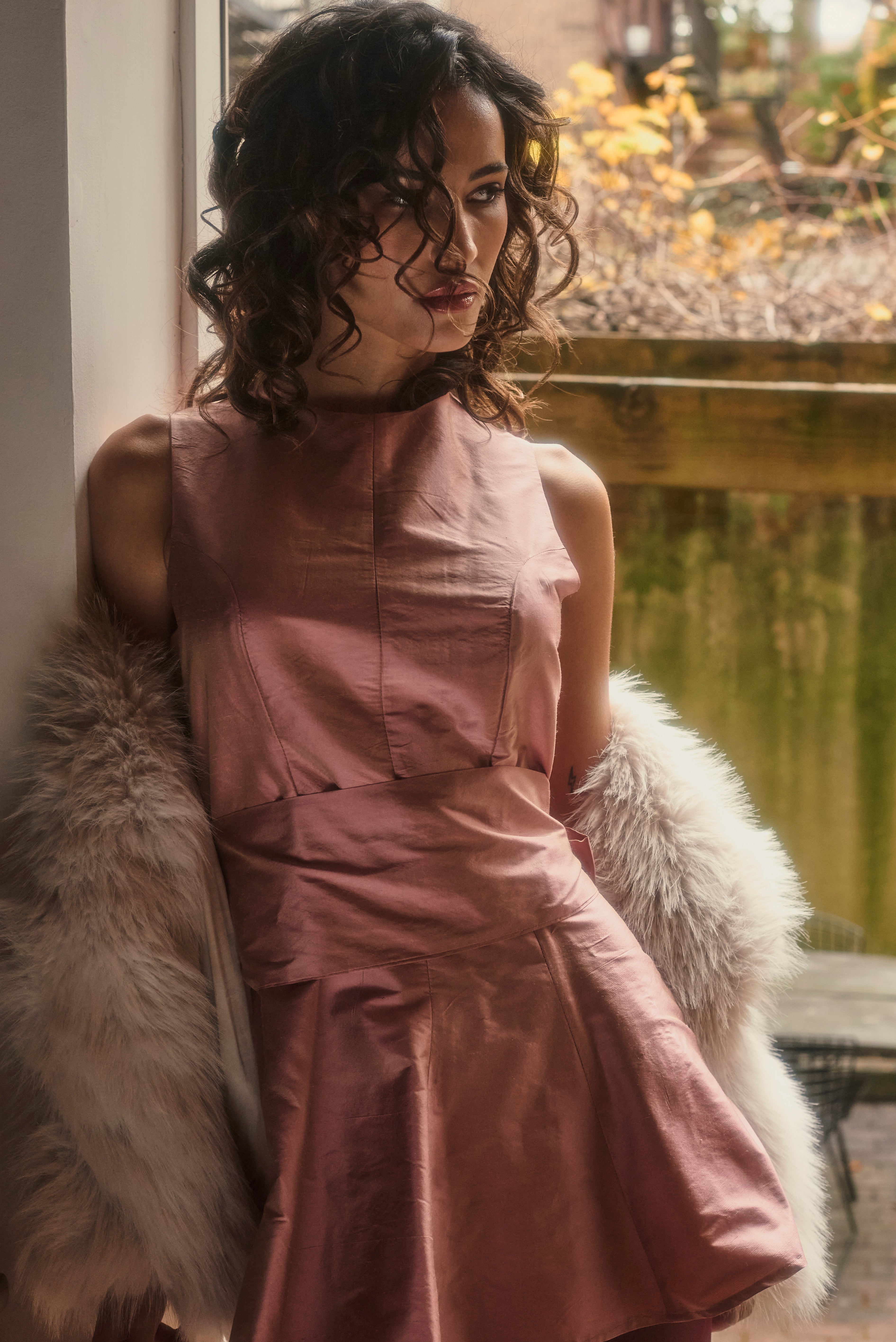 Woman in a pink dress sitting by a window with a view of a garden.