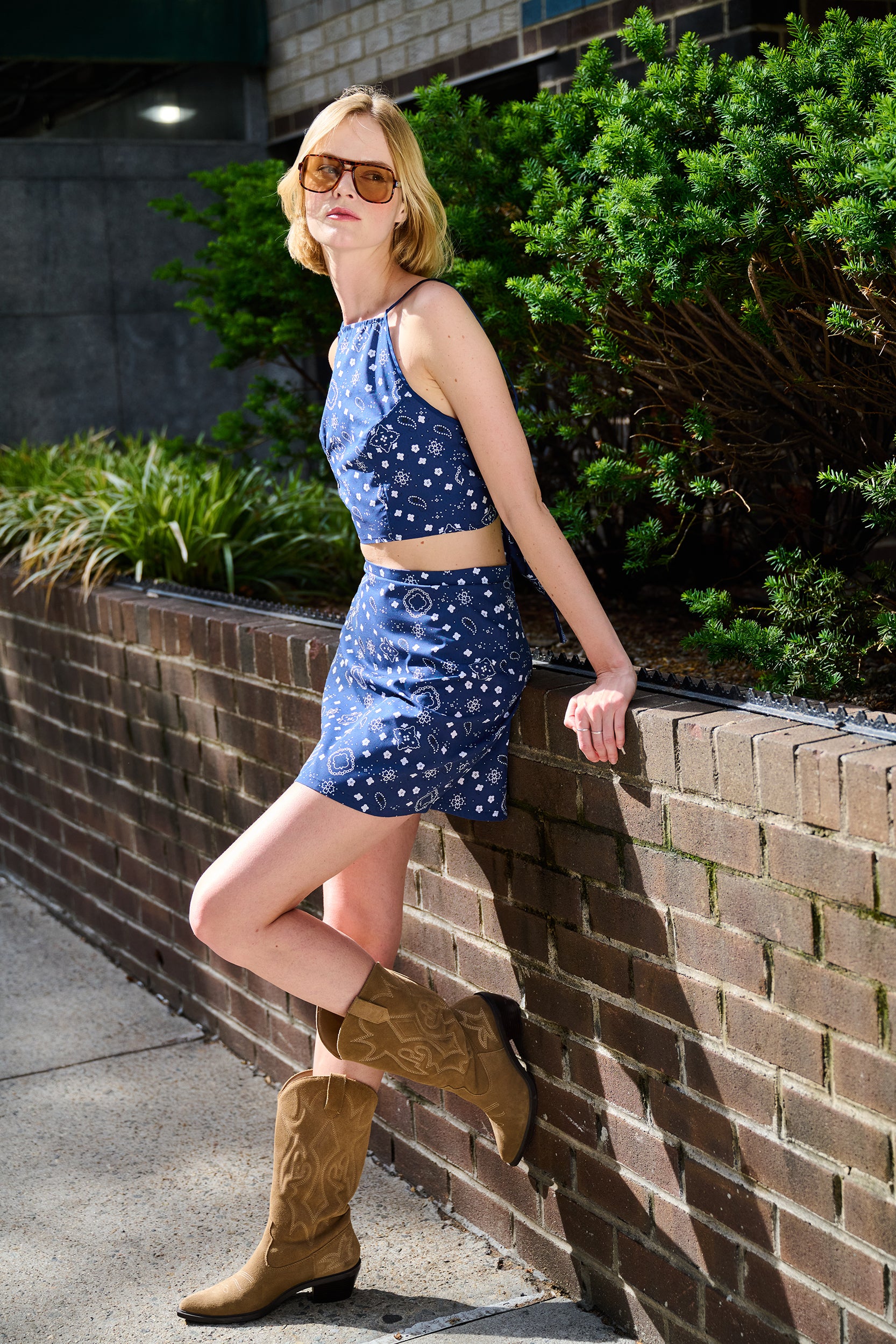 Woman in a blue navy bandana top and skirt set and brown boots leaning against a brick wall.