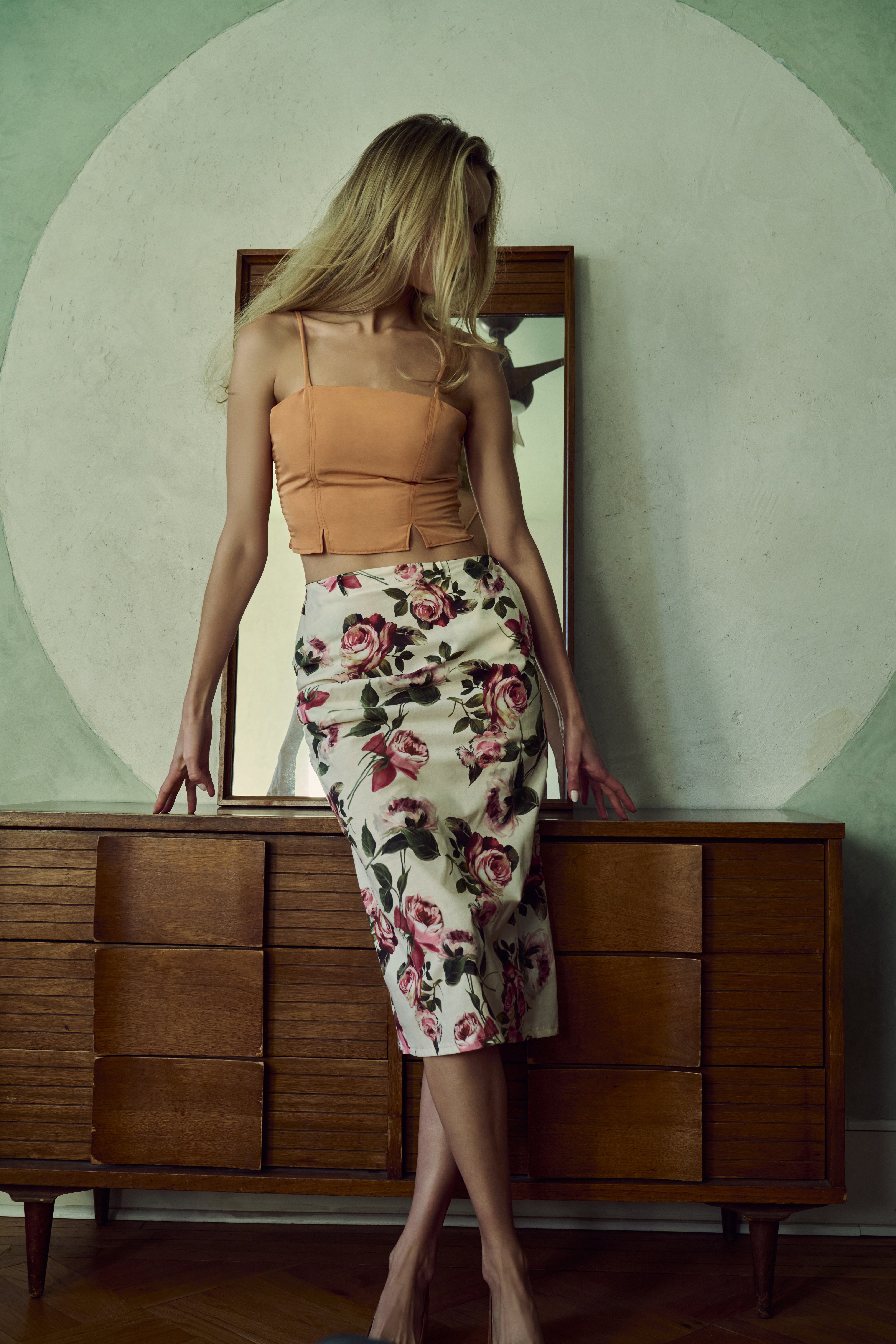 Woman in a floral skirt and orange top standing in front of a wooden dresser.